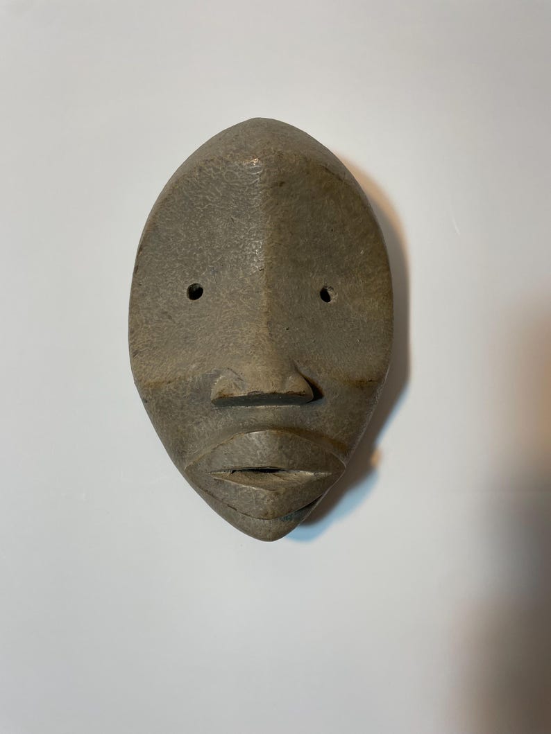 May include: A carved, grey wooden mask with a stylized face. The mask has a smooth, oval shape with two small eye holes, a prominent nose, and a closed mouth. The surface has a textured appearance.