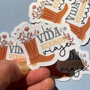 May include: A set of four stickers featuring a brown cowboy boot with floral accents and the text "La Vida es un Hermoso Viaje" in Spanish, which translates to "Life is a Beautiful Journey".