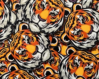 May include: Close-up of a pile of orange and black tiger face patches. The patches are made of fabric and have a stitched edge.