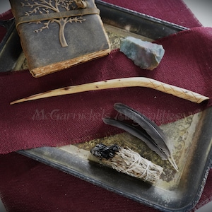 以下が含まれることがあります： 彫刻が施された木の杖、セージの束、黒い羽根2本、木製のデザインが施された革製のジャーナル、そしてグレーの石が、ワインレッドの布地の背景の上に置かれています。