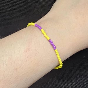 May include: A beaded bracelet featuring yellow and purple beads. The bracelet is made of small, round beads strung together to form a flexible band. The yellow beads make up the majority of the bracelet, with a section of purple beads in the center.