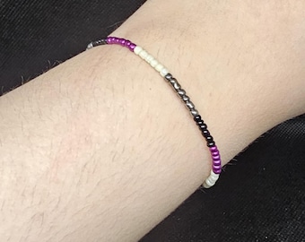 May include: A beaded bracelet featuring the colors of the asexual pride flag: black, gray, white, and purple. The bracelet is made of small, round beads strung together. The bracelet is worn on a wrist.