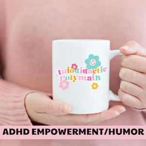 Może przedstawiać: Biały kubek ceramiczny z kolorową grafiką, na której widnieje napis "Autodidactic Polymath" z kwiatami i uśmiechniętymi buźkami.  ADHD Empowerment/Humor.