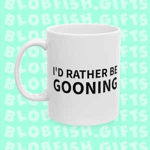 Puede incluir: Taza de cerámica blanca con asa curvada. La taza presenta el texto negro "I'D RATHER BE GOONING" centrado en la parte delantera. La taza es de tamaño estándar para café o té.