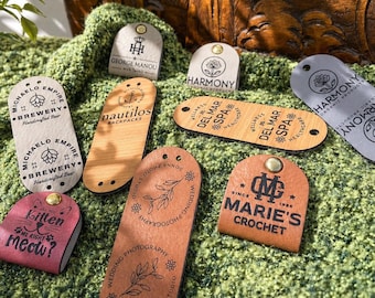 May include: A collection of various tags with different designs and text. The tags are made of wood, leather, and other materials. Some tags have logos, while others have text like "Kitten me right meow?" and "Marie's Crochet."