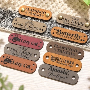 May include: A collection of rectangular, engraved leather tags in various colors, including brown, gray, and burgundy. The tags feature text such as "Flamingo to Tango," "Lazy Cat," and "Butterfly Cosmetics." Each tag has two holes and is secured with brass rivets.