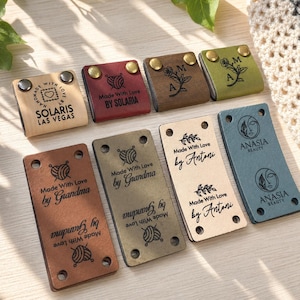 May include: A selection of leather labels in shades like tan, burgundy, and olive. The labels display text such as "Made With Love" and brand names, alongside designs like a yarn ball or floral motifs. Some labels are secured with metal rivets.