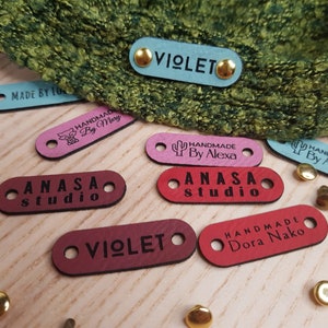 May include: Assortment of colorful leather labels with various text, including "Handmade by Alexa", "ANASA studio", "VIOLETO", and "Handmade Dora Nako". The labels are on a wooden surface with gold metal accents.