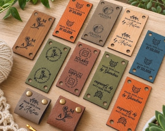 May include: A collection of leather tags in various colors, including brown, orange, and green. The tags feature embossed text and floral designs, with phrases like "Made With Love" and brand names. Some tags have metal rivets.