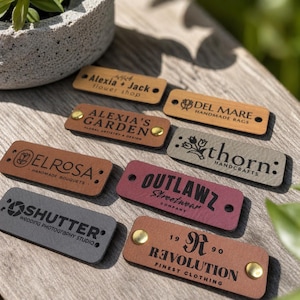 May include: A selection of rectangular leather tags in various colours, including brown, grey, and burgundy. Each tag features embossed text and designs, such as business names and logos. Some tags have brass rivets.
