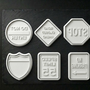 May include: A collection of white road sign-shaped cookie cutters. The shapes include a stop sign, a speed limit sign, a no parking sign, a road closed sign, a blank shield, and a do not enter sign.
