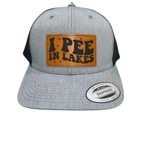 Puede incluir: Gorra de camionero gris y negra con un parche de cuero marrón. El parche muestra el texto "I PEE IN LAKES" en una fuente retro. La gorra tiene una visera curva y un cierre a presión.