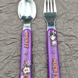 May include: A child's spoon and fork set featuring Mickey Mouse. The utensils have purple handles with the word "Mickey" printed in red and white. The spoon and fork are made of silver-colored metal.