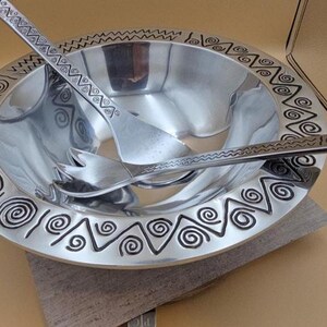 May include: A silver salad bowl with matching serving utensils. The bowl has a decorative border with spiral and zigzag patterns. The serving spoon and fork rest inside the bowl, ready for use. The bowl is sitting on a neutral-colored surface.