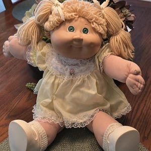 May include: A doll with blonde yarn hair styled in two pigtails tied with white ribbons, green eyes, and a soft fabric body. The doll wears a pale yellow dress with a white lace collar and trim, along with white fabric shoes and matching socks.