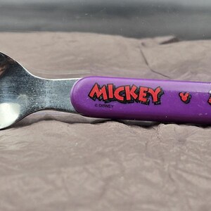 May include: A stainless steel spoon with a purple handle featuring the word "Mickey" in red and a graphic of Mickey Mouse. The spoon is designed for children and is likely part of a Disney-themed set.