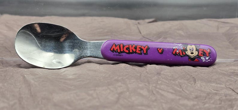 May include: A stainless steel spoon with a purple handle featuring the word "Mickey" in red and a graphic of Mickey Mouse. The spoon is designed for children and is likely part of a Disney-themed set.