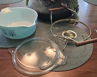 May include: A light blue glass casserole dish with a white floral pattern, a clear glass lid, and a gold-toned metal warming stand featuring two wooden handles. The dish has a pouring spout and the stand includes a central circular burner holder.