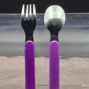 May include: A stainless steel fork and spoon set with purple handles. The fork has four tines, and the spoon has a rounded bowl. The handles are a solid, vibrant purple color.