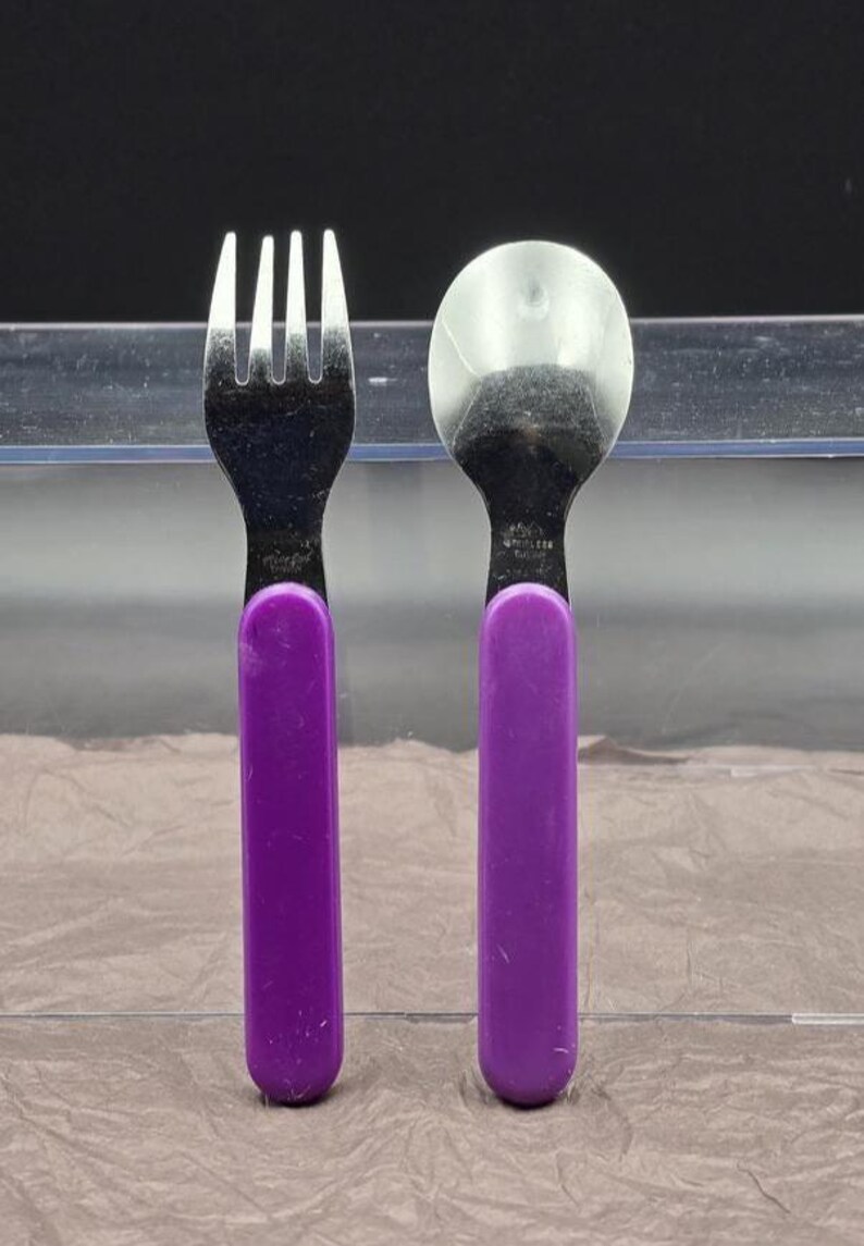 May include: A stainless steel fork and spoon set with purple handles. The fork has four tines, and the spoon has a rounded bowl. The handles are a solid, vibrant purple color.