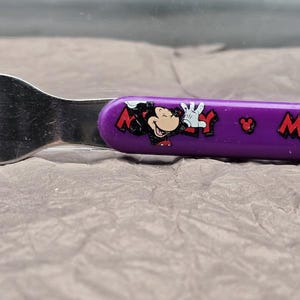 May include: A stainless steel fork with a purple handle featuring a Mickey Mouse graphic and the word "Mickey" in red. The fork has three tines and is designed for children's use.