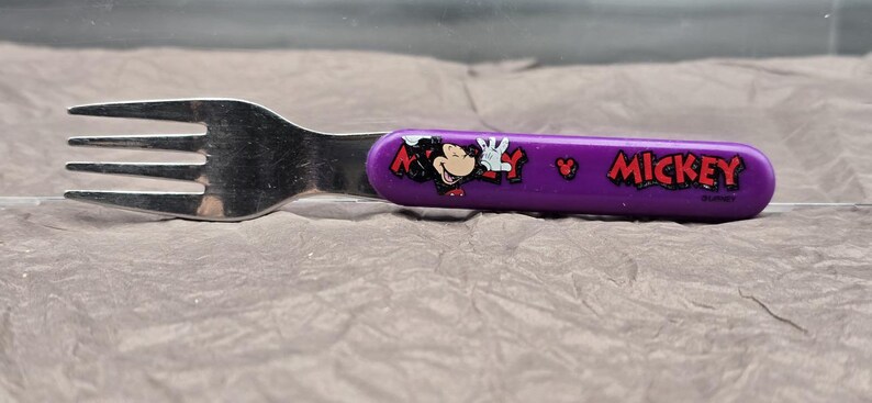 May include: A stainless steel fork with a purple handle featuring a Mickey Mouse graphic and the word "Mickey" in red. The fork has three tines and is designed for children's use.