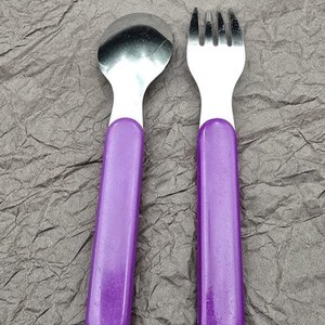 May include: A child's spoon and fork set. The spoon and fork have white tops and long, purple handles. The spoon has a rounded bowl, and the fork has four tines. The set is made of stainless steel and plastic.