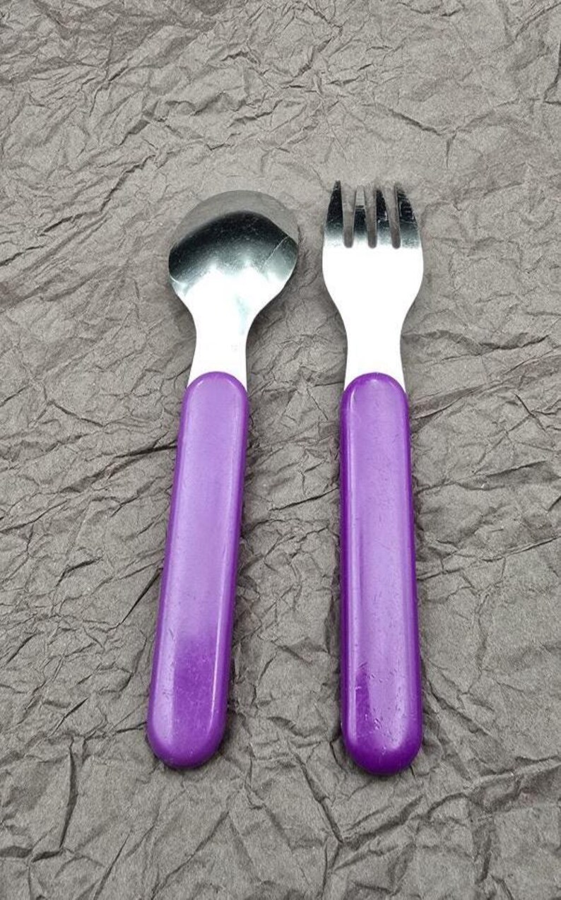 May include: A child's spoon and fork set. The spoon and fork have white tops and long, purple handles. The spoon has a rounded bowl, and the fork has four tines. The set is made of stainless steel and plastic.