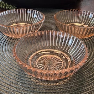 May include: Three small, round, light pink glass bowls featuring a vertical ribbed pattern along the exterior sides and a sunburst design on the interior base. The bowls have a scalloped edge and a translucent finish, reflecting light across the surface.