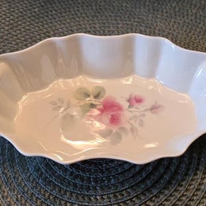 May include: An oval white ceramic dish with a scalloped, fluted rim. The center of the dish features a painted floral design consisting of a large pink rose, smaller rosebuds, and green leaves, all rendered in a soft, watercolor-like style.