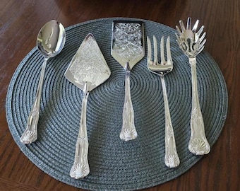 May include: A set of five silver-toned serving utensils including a large spoon, a triangular cake server, a rectangular flat server, a four-tined fork, and a pasta server. Each piece features a decorative shell motif at the base of the handle.