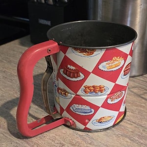 May include: A metal flour sifter with a red and white diamond pattern featuring illustrations of various baked goods. The sifter has a red handle with a squeeze-action lever mechanism and a dark interior rim, designed for kitchen baking tasks.