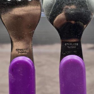 May include: Close-up of two stainless steel utensils: a fork and a spoon. Both have purple handles and are marked "STAINLESS TAIWAN". The utensils are likely part of a set of flatware.