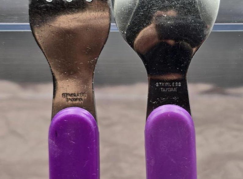 May include: Close-up of two stainless steel utensils: a fork and a spoon. Both have purple handles and are marked "STAINLESS TAIWAN". The utensils are likely part of a set of flatware.