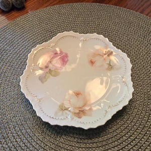 May include: A white porcelain plate with a scalloped edge and a gold-trimmed rim. The plate features three painted rose clusters in shades of soft pink, peach, and cream, arranged in a triangular pattern across the smooth, glossy surface.