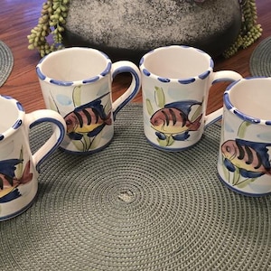 May include: A set of three white ceramic mugs featuring a hand-painted tropical fish design in shades of pink, yellow, and blue. Each mug has a scalloped rim with blue accents and a curved handle, showcasing a consistent aquatic theme throughout.
