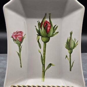 May include: A white rectangular ceramic tray with curved sides featuring a botanical illustration of three rose buds in varying stages of bloom, colored in shades of red and green, supported by a gold-toned twisted metal display stand.