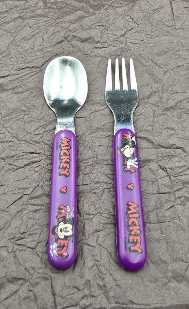May include: A child's spoon and fork set featuring Mickey Mouse. The utensils have purple handles with the word "Mickey" printed in red and white. The spoon and fork are made of silver-colored metal.