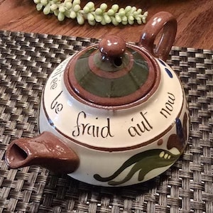May include: A ceramic teapot with a cream-colored base, featuring a brown handle, spout, and lid. The lid has a dark green center. The body is decorated with hand-painted green and blue floral motifs and black cursive text that reads fraid aut now.