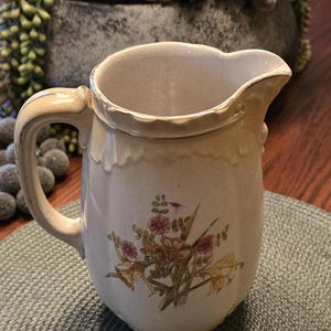 May include: A cream-colored ceramic pitcher featuring a delicate floral bouquet illustration on the front. The pitcher has a scalloped rim, a curved handle, and a subtle crackle glaze finish throughout the body, showcasing a classic design.