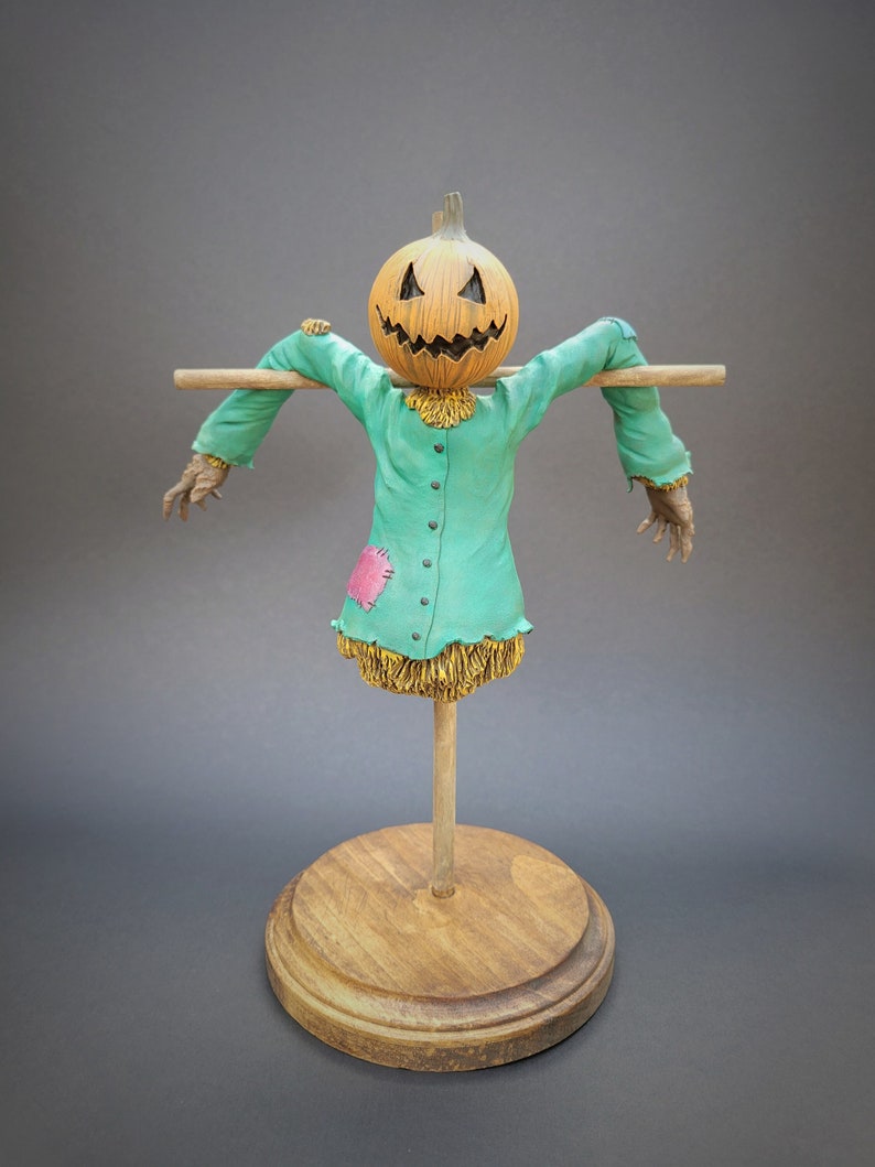 K&ouml;nnte beinhalten: Eine Vogelscheuchenfigur mit einem geschnitzten K&uuml;rbis-Kopf, die ein gr&uuml;nes Hemd mit einem rosa Fleck und stroh&auml;hnlichem Material am Boden tr&auml;gt. Die Vogelscheuche steht auf einem Holzst&auml;nder.