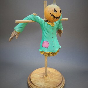 K&ouml;nnte beinhalten: Eine Vogelscheuchenfigur mit einem geschnitzten K&uuml;rbis-Kopf, die ein gr&uuml;nes Hemd mit einem rosa Patch und Stroh-Akzenten tr&auml;gt. Die Figur ist auf einem Holzst&auml;nder montiert.