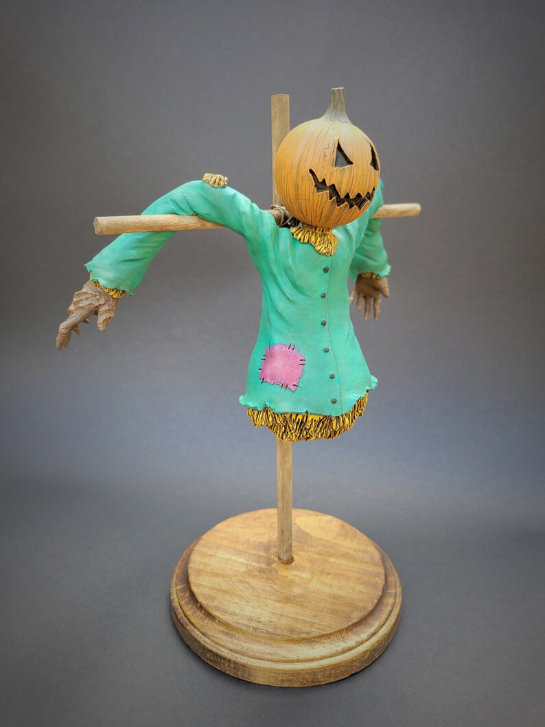 K&ouml;nnte beinhalten: Eine Vogelscheuchenfigur mit einem geschnitzten K&uuml;rbis-Kopf, die ein gr&uuml;nes Hemd mit einem rosa Patch und Stroh-Akzenten tr&auml;gt. Die Figur ist auf einem Holzst&auml;nder montiert.