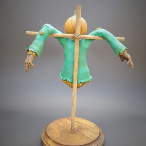 K&ouml;nnte beinhalten: Eine Vogelscheuchenfigur mit einem K&uuml;rbis-Kopf, einem gr&uuml;nen Hemd und strohhaltigen Armen und Beinen. Die Vogelscheuche ist auf einem Holzkreuz und einem Holzsockel montiert.