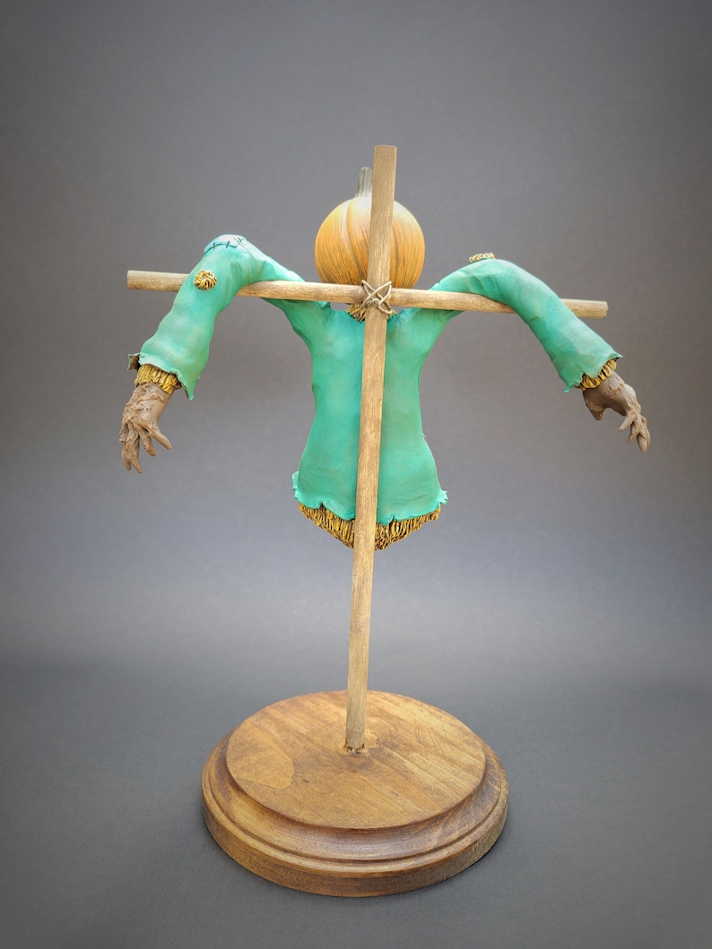K&ouml;nnte beinhalten: Eine Vogelscheuchenfigur mit einem K&uuml;rbis-Kopf, einem gr&uuml;nen Hemd und strohhaltigen Armen und Beinen. Die Vogelscheuche ist auf einem Holzkreuz und einem Holzsockel montiert.