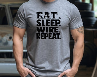 May include: A gray t-shirt with the text "EAT. SLEEP. WIRE. REPEAT." in black block letters. The shirt is a crew neck style and appears to be made of a soft material. The image is taken in a workshop setting.