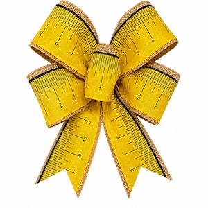 May include: A decorative bow made of yellow fabric with a black printed measuring tape pattern featuring numerical markings. The bow has a textured, natural-colored burlap-style edge along the perimeter of the ribbon loops and tails.