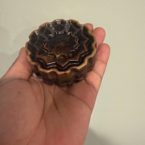 May include: A round, dark brown soap bar with a multi-layered, scalloped, or star-like edge design. The surface features a glossy finish with varying shades of brown, creating a marbled appearance across the concentric, tiered circular patterns.