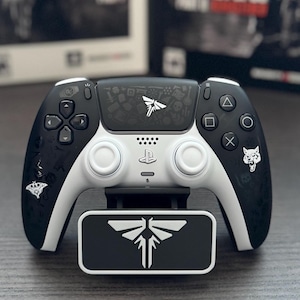 May include: A black and white PlayStation 5 controller with a matching stand, featuring a Firefly logo and The Last of Us branding. Two game boxes are in the background.
