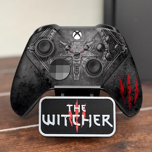 May include: A black Xbox controller with a Witcher-themed design, featuring a wolf emblem and red claw marks. The controller rests on a stand with "THE WITCHER" text. A black console is visible in the background.